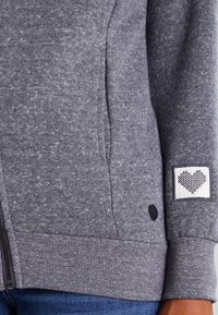 Felpa grigia con zip realizzata in tessuto testurizzato. Presenta una patch a forma di cuore sulla manica e un'etichetta con il logo vicino all'orlo.