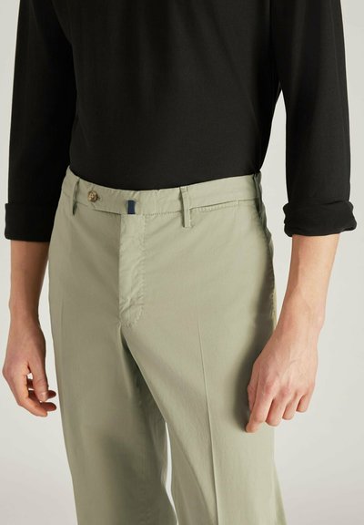 Lichtgroene broek van een gladde stof, met een rits- en knoopsluiting, voorzakken en opgerolde mouwen van een zwart shirt.