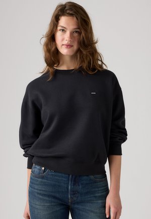 Jeune femme aux cheveux bruns ondulés portant un sweat-shirt noir Levi’s et un jean bleu sur un fond gris uni.