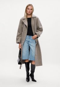 Abrigo de lana gris claro con cuello de chal, top negro ajustado, culottes de mezclilla azul hasta la rodilla y botas negras de puntera afilada. Lleva un bolso negro.