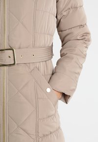 Světle béžový prošívaný péřový kabát s páskem v pase, zlatým zipem a dvěma postranními kapsami se zapínáním na patentky.