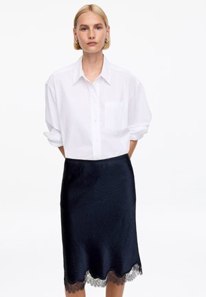 Kobieta w białej koszuli z guzikami i podwiniętymi rękawami oraz w czarnej spódnicy do kolan z koronkowym wykończeniem, stojąca z rękami za plecami.