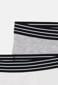 Două perechi de lenjerie pentru bărbați cu designuri contrastante ale chingilor taliei; una gri cu o bandă neagră și albă cu dungi și una albă cu o bandă similară.