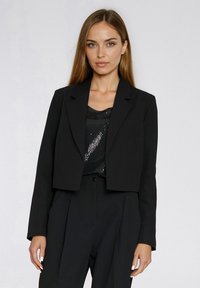 Przycięta czarna marynarka z klapą z wycięciem, gładką teksturą i prostym krojem, nałożona na czarny top z cekinów i pasujące spodnie.