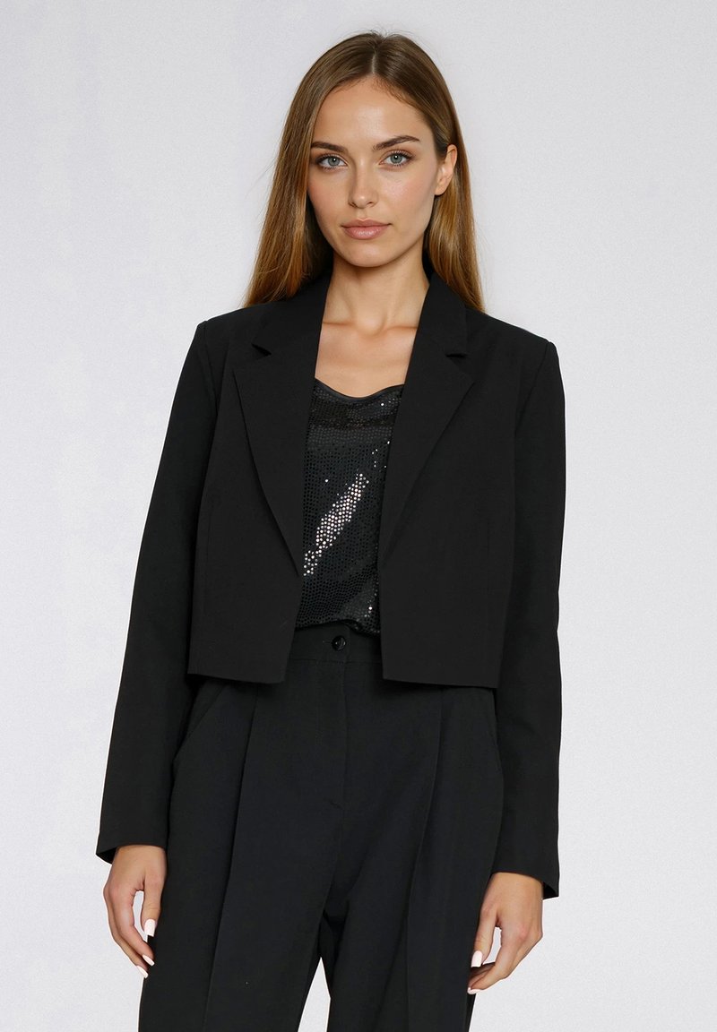 Przycięta czarna marynarka z klapą z wycięciem, gładką teksturą i prostym krojem, nałożona na czarny top z cekinów i pasujące spodnie.