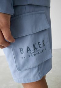 Ljusblå bomullsshorts med en stor, platt ficka som har en tryckt logotyp med texten "BAKER LONDON BY TED BAKER." Slät yta.
