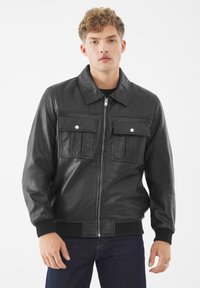 Junger Mann in einer schwarzen Lederjacke mit Reißverschluss und zwei Klappentaschen auf der Brust, kombiniert mit dunkelblauen Jeans, steht vor einem weißen Hintergrund.