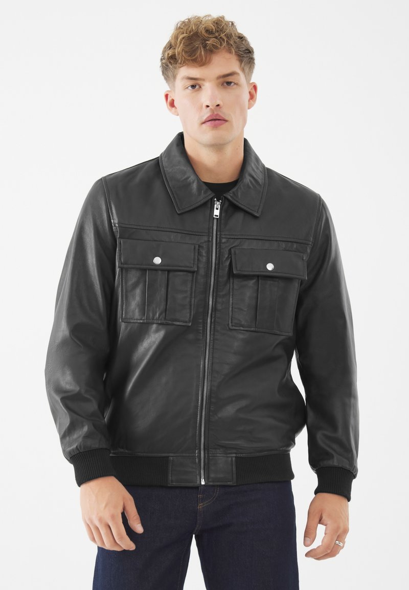 Junger Mann in einer schwarzen Lederjacke mit Reißverschluss und zwei Klappentaschen auf der Brust, kombiniert mit dunkelblauen Jeans, steht vor einem weißen Hintergrund.