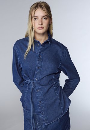 Giacca in denim di colore blu medio con chiusura a bottoni, maniche lunghe e vita con cordoncino per una vestibilità regolabile. Presenta cuciture a contrasto.