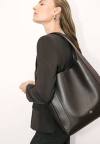 Brun lædertaske med en glat tekstur, minimalistisk design og en enkelt skulderrem. Har diskret logo foran.