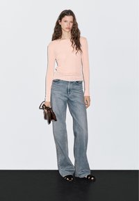 Langärmliges, hellrosa Oberteil kombiniert mit weit geschnittenen, hellblauen Jeans. Das Model trägt eine braune Handtasche und schwarze Sandalen. Einfarbiger Hintergrund.