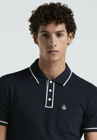 Polo-shirt bleu marine avec un col et une patte de boutonnage blancs contrastants, un petit poche poitrine et un détail de logo. Texture de tissu douce.