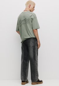 Tricou oversized de culoare verde măsliniu cu logo brodat pe spate, asortat cu blugi negri cu picioare largi și pantofi cu platformă maronii.