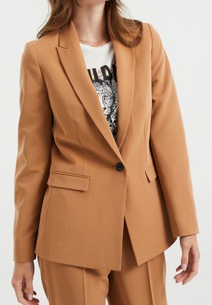 Ljusbrun skräddarsydd blazer med slag, enkel knappstängning och sidofickor. Bärs över en vit grafisk t-shirt av strukturerad tyg.