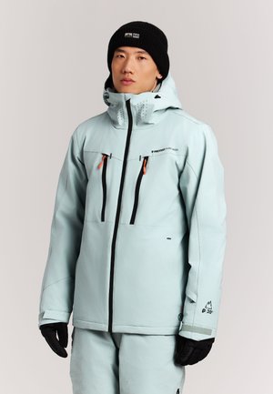 Jonge man draagt een lichtblauwe winterjas met ritssluitingen op de borstzakken, zwarte handschoenen en een zwarte muts tegen een effen achtergrond.