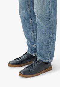 Mornarski usnjeni superge z rjavimi gumijastimi podplati, z vezalnim dizajnom in teksturiranimi detajli, nosijo jih z svetlo modrimi jeans hlačami.