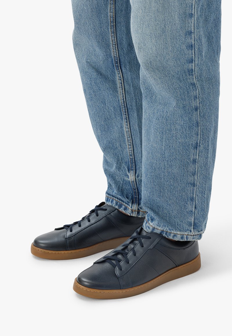 Mornarski usnjeni superge z rjavimi gumijastimi podplati, z vezalnim dizajnom in teksturiranimi detajli, nosijo jih z svetlo modrimi jeans hlačami.