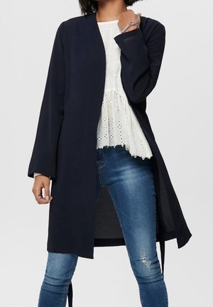 Manteau long bleu marine en tissu doux, avec une coupe droite et des manches larges, associé à un haut en dentelle blanche et un jean bleu déchiré.