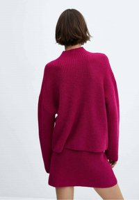 Fuksjowy sweter z dzianiny o wysokim kołnierzu i luźnym kroju, zestawiony z dopasowaną spódnicą ze żebrowanym wzorem. Obie rzeczy mają teksturowane wykończenie.