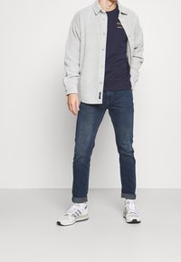Camisa de pana gris claro con botones, sudadera azul marino de cuello redondo debajo, jeans azul oscuro y zapatillas grises con acentos en blanco y negro.