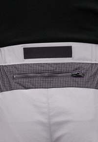 Φωτεινό γκρι παντελόνι με οριζόντια τσέπη σε μοτίβο μαύρου πλέγματος και μαύρο ιμάντα βέλκρο πάνω από τη ζώνη, σε φόντο μαύρου τοπ.