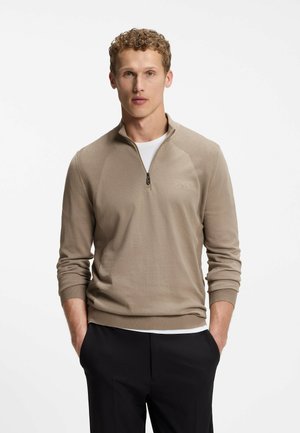 Homme aux cheveux bouclés portant un pull beige à demi-fermeture éclair sur une chemise blanche et un pantalon noir, debout les mains dans les poches sur fond blanc.