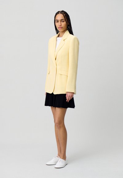 Blazer oversize giallo chiaro con spalle strutturate, due tasche frontali e chiusura con un bottone, abbinato a una gonna plissettata nera e scarpe bianche.
