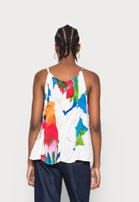 Bílý top bez rukávů s květinovým vzorem, který obsahuje jasné barvy jako červená, modrá a zelená. Lehce se nosící látka s volným střihem.