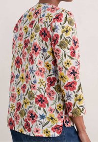 Camicia bianca con una stampa floreale colorata che presenta fiori rossi, rosa, gialli e blu; maniche a tre quarti; spacco ai lati dell'orlo.