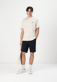 Lys beige bomulls t-skjorte med en gul logo, parret med marineblå shorts, hvite joggesko og en beige totebag. Enkel design og uformell passform.