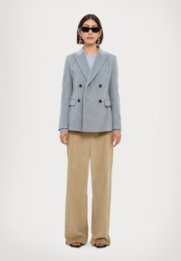 Blazer bleu clair croisé avec des boutons noirs, associé à un pantalon en velours côtelé beige à jambes larges. Le mannequin porte des lunettes de soleil noires et de grosses boucles d'oreilles.