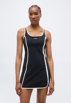 Sieviete ar pīti matu sānu sēž, tērpusies melnā bezroku kleitā ar baltu apmali un "GUESS" logotipu, stāvot pret vienkāršu gaišu fonu.