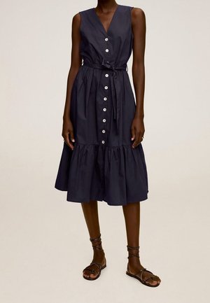 Robe midi sans manches bleu marine à boutons, avec ceinture à la taille, portée avec des sandales plates noires à lanières sur une personne debout devant un fond uni.
