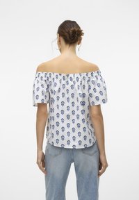 Hvid off-shoulder bluse med blåt blomsterdesign, korte pufærmer og elastisk halsudskæring, parret med lyse denimjeans.