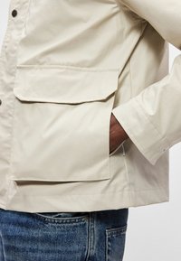 Μπεζ αδιάβροχο jacket με μεγάλη τσέπη με καπάκι, κουμπιά με ασφάλεια και λεία υφή, σε συνδυασμό με μπλε τζιν παντελόνι.