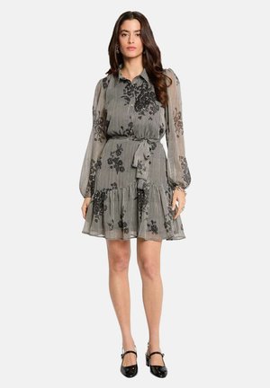 Femme portant une robe transparente à manches longues cintrée à la taille, avec des motifs floraux noirs sur de fines rayures, associée à des chaussures Mary Jane noires.