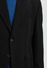 Černý texturovaný blazer se dvěma knoflíky přes modrý pletený svršek, s náprsní kapsou a klopovou kapsou na pravé straně.