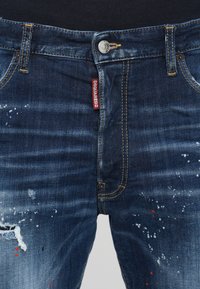 Blugi din denim bleumarin închis, cu detalii uzate, decolorare ușoară, stropi de vopsea albă și roșie și eticheta DSQUARED2 pe talie.