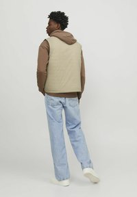 Gilet beige clair porté sur un sweat à capuche marron, associé à un jean bleu clair. Le modèle est vu de dos, mettant en avant une coupe décontractée et un style casual.
