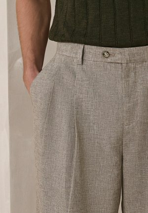 Mand iført lyse beige teksturerede bukser med knaplukning og en mørkegrøn ribstrikket skjorte, hånd i lommen.