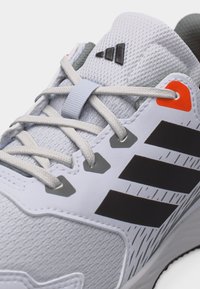 Λευκό και γκρι αθλητικό παπούτσι Adidas με μαύρες τρεις ρίγες, λευκά κορδόνια και πορτοκαλί λεπτομέρεια κοντά στο επάνω αυτί.