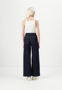 Široke temnodenske jeans hlače s kontrastnim šivanjem, visokim pasom in dvema zadnjima žepoma, v kombinaciji z belo majico brez rokavov in športnimi copati.