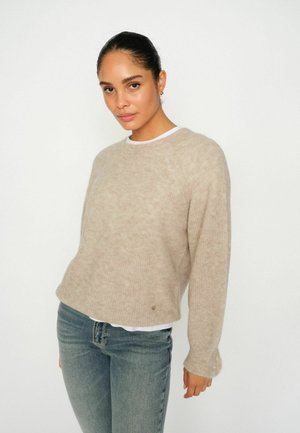 Beige knitted sweater with a relaxed fit, round neckline, and ribbed cuffs, layered over a white shirt, worn with light blue jeans.