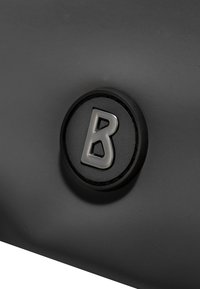 Črno teksturirano površje s krožnim logotipom, ki vsebuje veliko črko "B" v srebrni barvi. Logotip je centriran v dvignjenem krožnem delu.