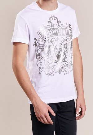 T-shirt en coton blanc avec un motif graphique argenté orné de motifs élaborés et d'un lion, assorti à un pantalon noir.
