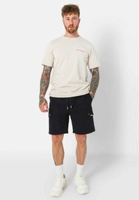 T-shirt beige in cotone a maniche corte con testo rosa, abbinato a pantaloni cargo scuri con tasche laterali. Sneakers bianche e calzini alla caviglia in vista.
