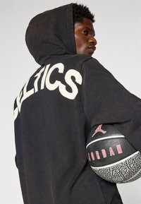 Sort hættetrøje med "CELTICS" i hvide bogstaver på ryggen, parret med en sort basketball mærket med Jordan-branding og tekstureret overflade.