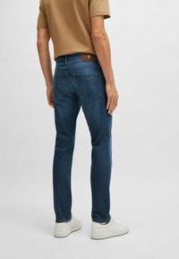 Jean en denim bleu foncé avec une coupe slim, comportant quatre poches, des coutures contrastées et une étiquette en cuir marron au niveau de la ceinture.