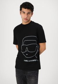 Black cotton t-shirt featuring a white line drawing of a face wearing sunglasses, with "KARL LAGERFELD" text below. Classic crew neck design.