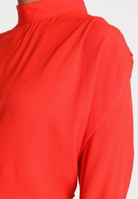 Blouse rouge à col montant avec manches longues. Fabriquée en tissu texturé. Présente une coupe ample avec fronces au niveau du décolleté.